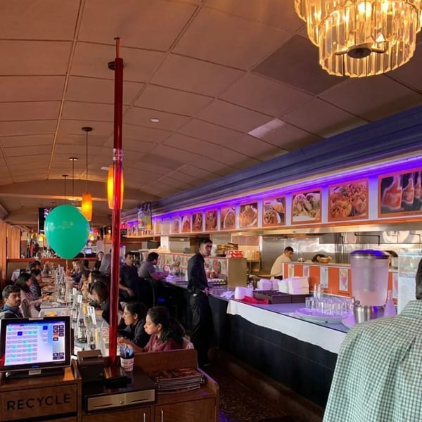 Bustling diner interior with customers seated at a long counter, staff, and food menu displays.