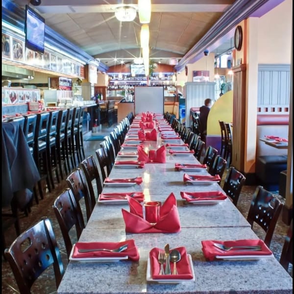 Long restaurant dining table set with red napkins and cutlery, with a bar and booths visible.