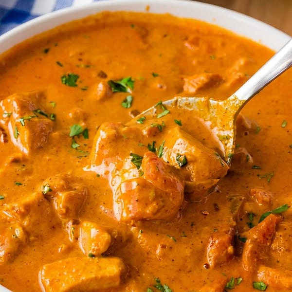 A white bowl of chicken tikka masala with cilantro, a spoon, and a blue checkered napkin.